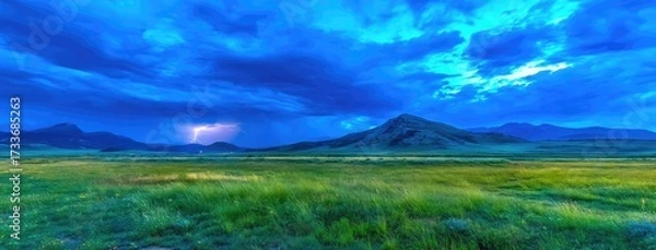 Fototapeta Dramatic lightning streaks across a dark blue sky over a tranquil mountain landscape at dusk, casting a vibrant glow over the lush green meadow