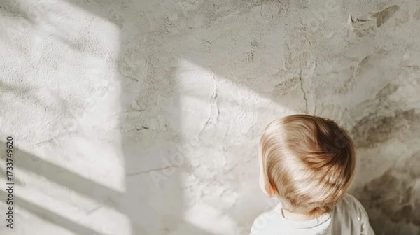 Fototapeta A baby is looking at the camera with a curious expression. The room is plain and white, with a window in the background. The baby's head is the main focus of the image