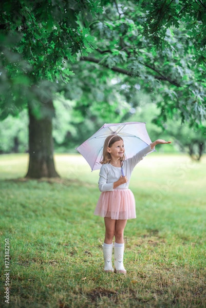 Fototapeta Little girl smiling while raising one hand to feel the rain under her umbrella. A joyful and curious moment of sensory exploration in a rainy park.
