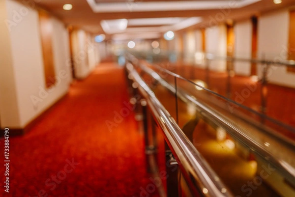 Fototapeta Modern hotel hallway with red carpet, warm ambient lighting, and sleek glass-metal railings in focus, creating a clean and stylish interior perspective.