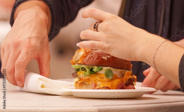 Fototapeta Two people at a food festival in Karlín Square, Prague, sharing a freshly prepared gourmet burger with melted cheese and vegetables, highlighting a social dining moment.