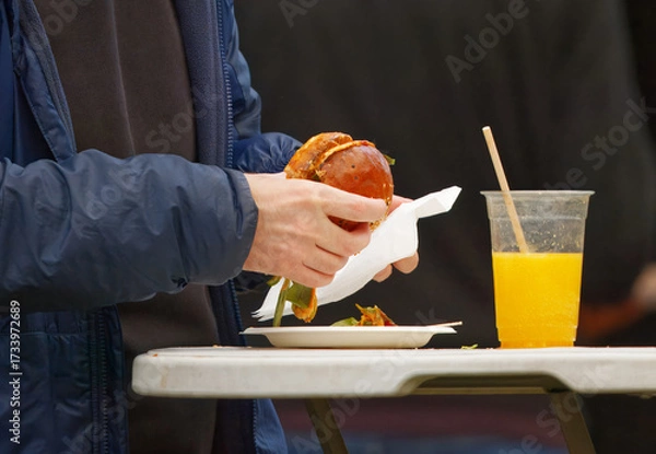 Obraz Midsection view of a visitor enjoying a juicy burger with a glass of orange juice at a food festival in Karlin, Prague, authentic street food atmosphere.