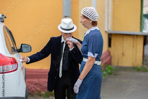 Fototapeta An elegant elderly couple, engaged in a romantic gesture near a vintage car in a charming city atmosphere in the daytime