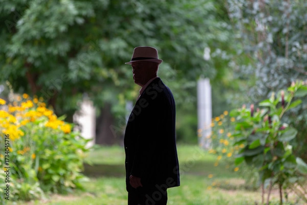 Fototapeta Elegant man in a hat stands in silhouette against a vibrant garden backdrop on a sunny day
