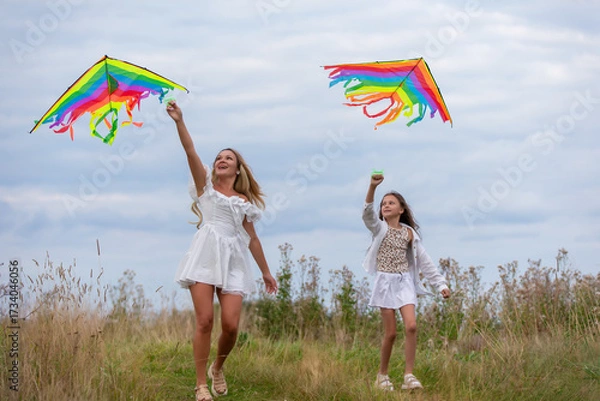 Fototapeta Two girls joyfully flying colorful kites in a grassy field under a cloudy sky during a pleasant afternoon