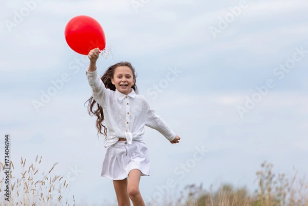 Fototapeta Young girl runs joyfully in a field holding a bright red balloon on a cloudy day