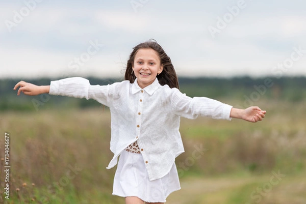 Fototapeta Smiling girl in white outfit joyfully running through a green field on a cloudy day