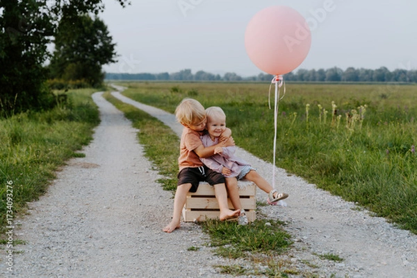 Fototapeta Brother and sister sitting together on wooden box in countryside. Happy siblings in nature. Portrait of little girl and boy. Children birthday party, outdoor nature photoshoot with festive atmosphere.