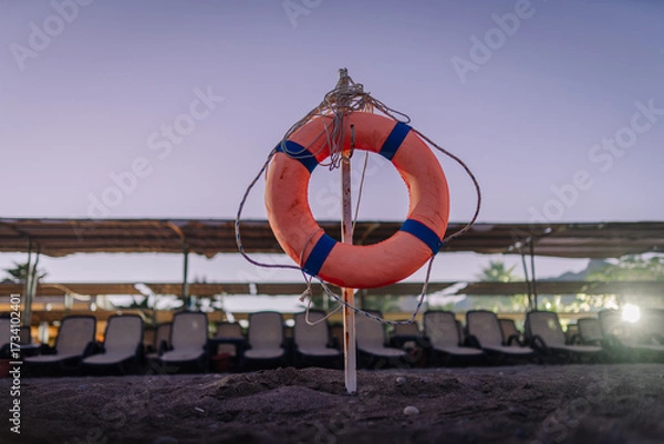 Fototapeta A lifebuoy ring mounted on a pole in the sand at a beach with empty loungers and canopies in the background during dusk.