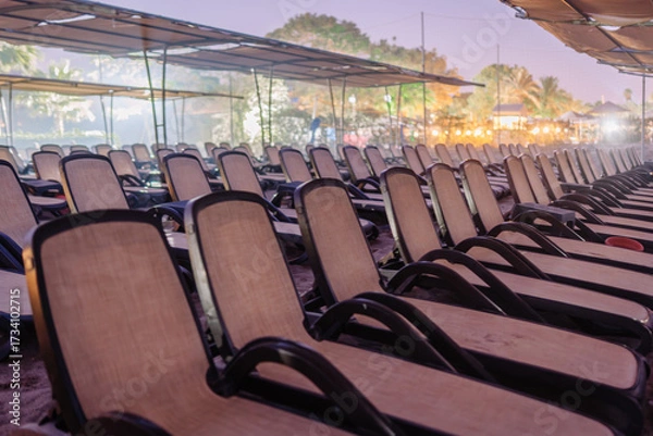 Fototapeta Rows of empty beach loungers arranged under canopy shades on a sandy beach at dusk, with soft lighting and a relaxed resort ambiance.