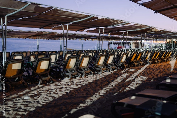 Fototapeta Rows of empty sun loungers under canopies on a sandy beach at dusk, facing the calm sea with city lights glowing in the distance.