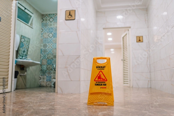 Fototapeta Yellow caution sign reading “Cleaning in Progress” placed in a tiled public restroom hallway with visible sinks and accessible restroom signs.