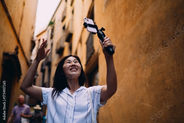 Fototapeta Happy female content creator records joyful video on smartphone stabilizer, embracing real-time sharing and online storytelling while walking confidently in sunlit city street.