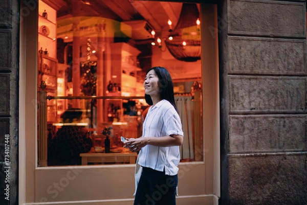 Fototapeta Asian woman stands outside warmly lit cafe, holding phone in hand, embracing peaceful digital pause during an urban stroll and reflecting quiet joy in everyday connectivity.