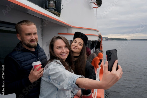 Fototapeta Three people are on a boat on the water one woman is taking a selfie with her phone. The other two are beside her. It seems they are having fun