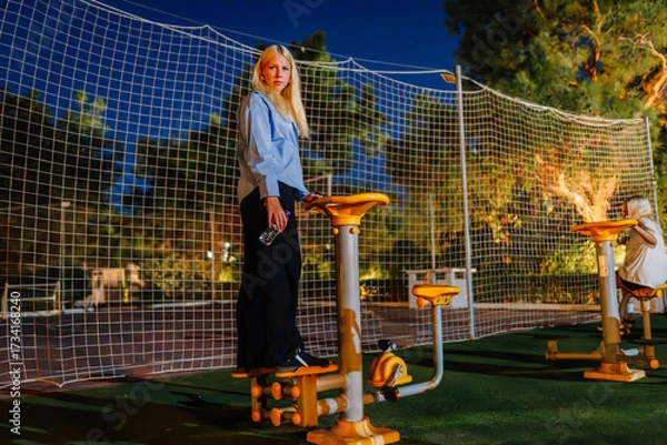 Fototapeta A woman uses outdoor gym equipment at night, standing on an exercise machine while holding a water bottle, with a net and trees in the background.