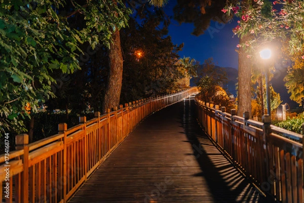 Fototapeta Wooden boardwalk path surrounded by trees, illuminated at night with warm lights, creating a peaceful and scenic atmosphere in a park or forest area.