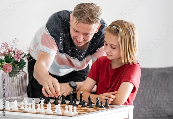 Fototapeta Thoughtful, Cute Little Girl Plays Chess With Father As He Explains How The Pieces Move