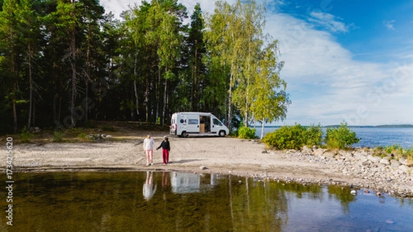Fototapeta Two friends stroll along the sandy shore of Lake Saimaa, surrounded by lush greenery and a tranquil atmosphere. A camper van is parked nearby, ready for exploration.