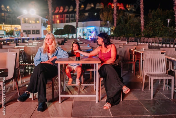 Fototapeta Three females, including a child, sit at an outdoor table at night. One woman reaches toward the child, all lit by colorful event lighting in a relaxed setting.