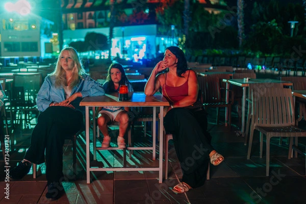 Fototapeta Three females, including a child, are seated at a table in an outdoor venue at night, illuminated by colorful lights, watching something off-camera.