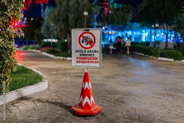 Fototapeta A bright warning sign on a traffic cone prohibits electric vehicles, displayed in three languages, placed on a cobblestone path in a park at night.