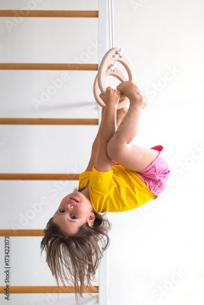 Fototapeta Little girl child age 3 4 years old in yellow shirt and pink shorts hanging upside down on gymnastic rings at home showing strength flexibility and playful childhood activity, vertical