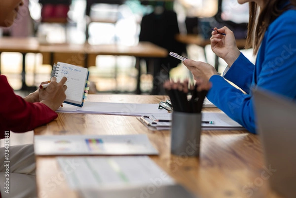 Fototapeta Businesswoman having meeting taking notes using smartphone in modern office. Two businesswomen in a bright office, discussing plans and taking notes on a smartphone.
