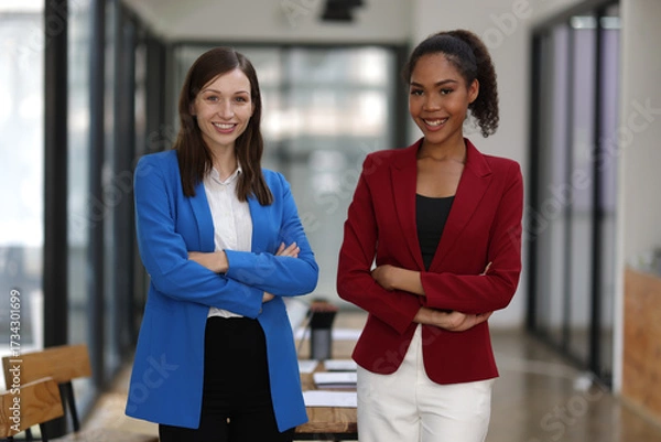 Fototapeta Businesswoman smiling with arms crossed in office. Two confident businesswomen in a modern office, arms crossed, wearing colorful suit, showing teamwork and success.