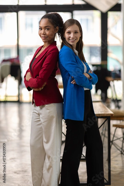 Fototapeta Businesswoman smiling with arms crossed in office. Two confident businesswomen in a modern office, arms crossed, wearing colorful suit, showing teamwork and success.