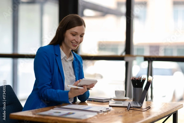 Fototapeta Businesswoman taking notes while having video call on laptop in office. Confident woman smiling during video call in modern office, taking notes. Professionalism and dedication in remote work era.