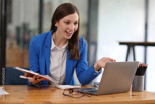 Fototapeta Businesswoman taking notes while having video call on laptop in office. Confident woman smiling during video call in modern office, taking notes. Professionalism and dedication in remote work era.