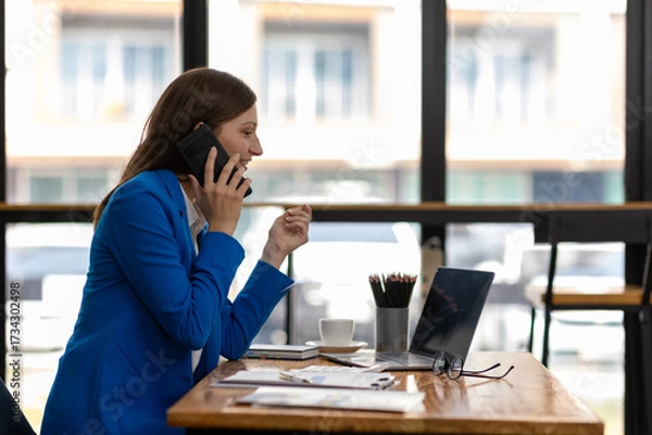 Fototapeta Businesswoman working from home office and talking on phone. She chatting on the phone and glancing at her laptop screen.