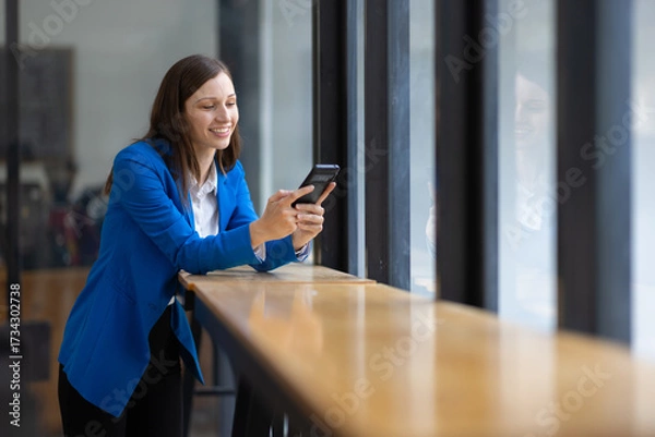 Fototapeta Businesswoman using smartphone and looking through office window. Happy woman taking a break in a modern office, using her smartphone and looking out the window.