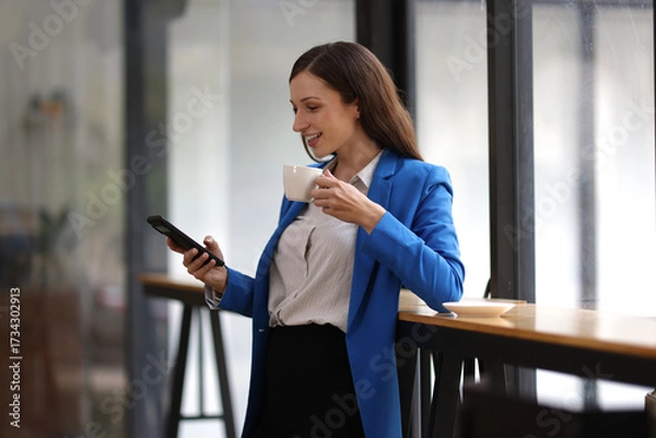 Fototapeta Businesswoman using smartphone and looking through office window. Happy woman taking a break in a modern office, using her smartphone and looking out the window.