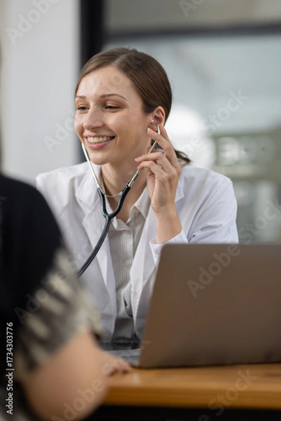 Fototapeta Doctor examining patient using stethoscope in hospital office. Female doctor listens to a patient's heartbeat with stethoscope during medical exam. She focuses on heart and lung health, care expertise