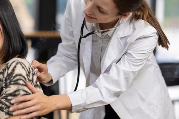 Fototapeta Doctor using stethoscope listening patient back during medical checkup. Doctor wearing white coat is examining a female patient using stethoscope, pressing it against her back to listen to her lungs.