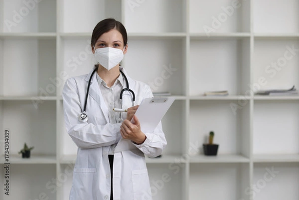 Fototapeta Confident female doctor in a modern clinic, holding a clipboard and wearing a protective mask, conveys reassurance and dedication, providing professional care in a bright and clean office setting.