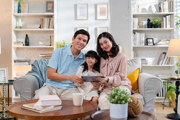 Fototapeta Happy Asian family using tablet,laptop for video call,playing game in the living room and looking at the camera.Smiling parents,their daughter are using a digital tablet for learning or entertainment
