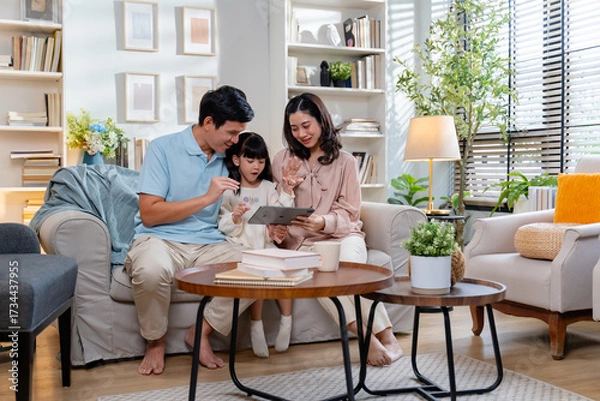 Fototapeta Happy Asian family using tablet,laptop for video call,playing game in the living room and looking at the camera.Smiling parents,their daughter are using a digital tablet for learning or entertainment