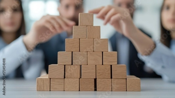 Fototapeta A group of professionals collaboratively builds a wooden block pyramid, symbolizing teamwork and strategy in a modern office setting.