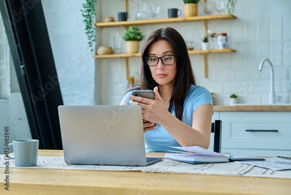 Fototapeta Young woman working with laptop, using smartphone sitting at table at home