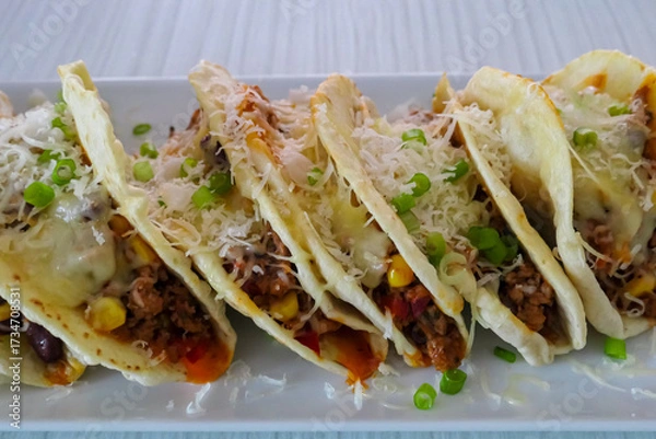 Obraz A close-up of delicious homemade tacos on a white plate. Each tortilla is filled with seasoned ground meat, corn, and beans, then generously topped with melted cheese and fresh chopped green onions.