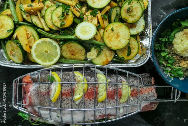Obraz A whole raw trout seasoned with herbs and lemon slices rests in a grilling basket next to a pan of fresh vegetables, all prepared and ready for a delicious and healthy summer barbecue cookout at home.