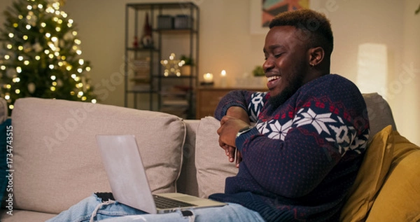 Obraz On a winter evening, a cheerful young student of multi-ethnic background sits on a couch, watching a funny video from yesterday's party. The young man laughs sincerely and is in a good mood. He wears