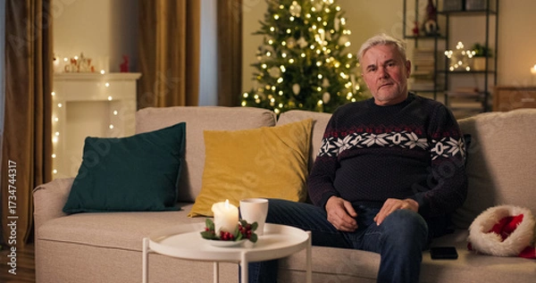 Fototapeta Sitting on a sofa in the living room on Christmas Eve, a lonely middle-aged man dropped his Santa's hat in disappointment. Left alone today, he set the hat aside and wears a warm sweater.