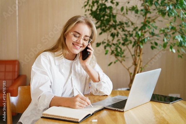 Fototapeta Young woman working on laptop while talking on phone and taking notes