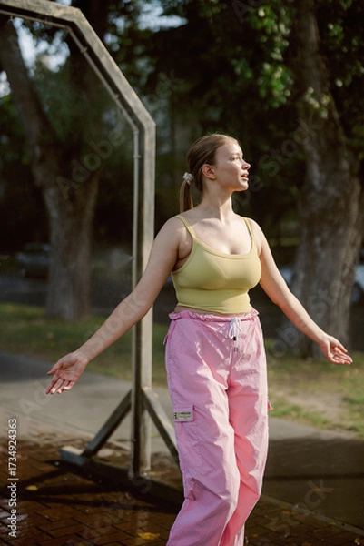 Fototapeta A woman stands in a park, soaking up the sun while wearing a yellow top and pink pants. She smiles and spreads her arms, enjoying a joyful moment in nature on a warm day.