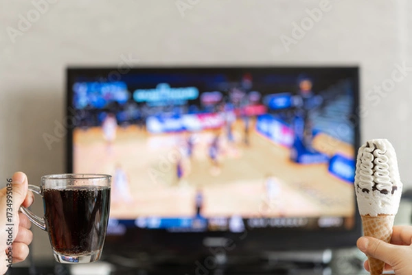 Fototapeta hands holding cup of cola and ice cream cone with a TV broadcasting basketball match on background