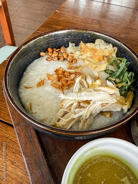 Fototapeta Traditional Indonesian chicken porridge or Bubur Ayam served with crackers, chili sauce, green sauce, and toppings on a wooden tray over rustic wooden table.